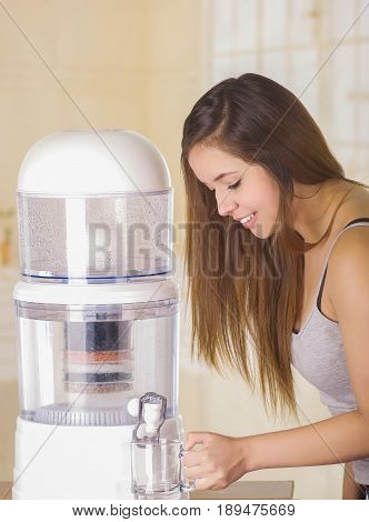 Beautiful smiling woman filling a glass of water, with a filter system of water purifier on a kitchen background.