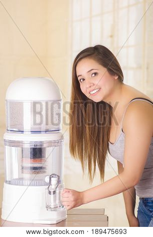 Beautiful smiling woman filling a glass of water, with a filter system of water purifier on a kitchen background.