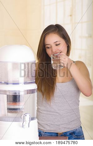 Beautiful woman drinking a glass of water with a filter system of water purifier on a kitchen background.