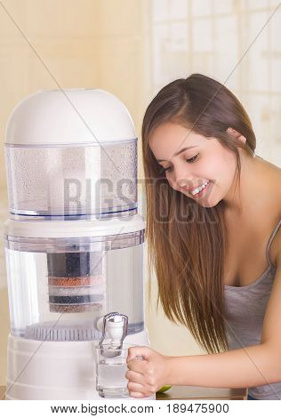 Beautiful smiling woman filling a glass of water, with a filter system of water purifier on a kitchen background.