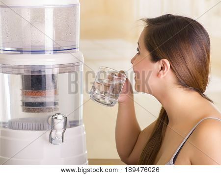 Close up of a beautiful woman drinking a glass of water with a filter system of water purifier on a kitchen background.