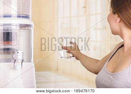 Beautiful smiling woman holding a glass of water, with a filter system of water purifier on a kitchen background.