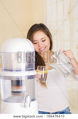Beautiful woman holding a glass of water in one hand and a pitcher of water in her other hand, with a filter system of water purifier on a kitchen background.