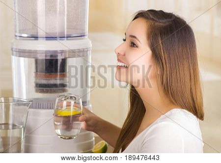 Beautiful smiling woman holding a cut lemon in her hand with a filter system of water purifier on a kitchen background.