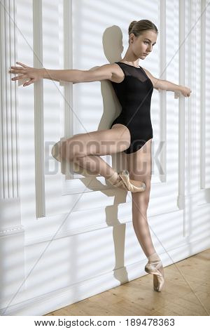 Charming ballerina leans with stretched arms on the white wall while stands on the left toe. She wears a black leotard with pointe shoes and looks forward. Stripes shadows are on the wall. Indoors.