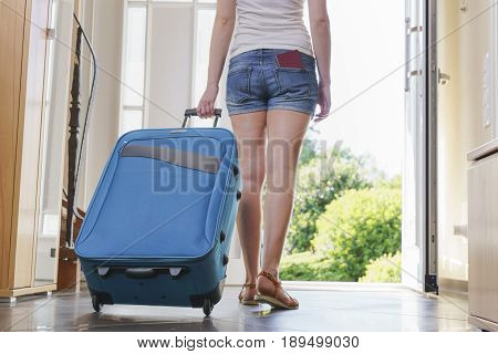 young woman In T-shirt, shorts and sandals with blue vacation bag exits through the front door.