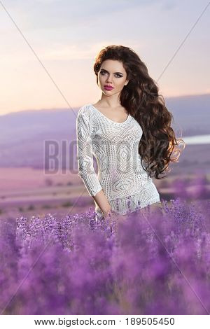 Beautiful carefree young woman portrait in lavender field at sunset. Attractive brunette girl with healthy long curly hair style enjoying life over nature landscape background.
