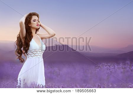 Enjoyment. Wellness. Beautiful brunette in lavender field. Carefree young woman portrait  over ridge mountains at sunset. Attractive girl with healthy long curly hair enjoying life on nature.