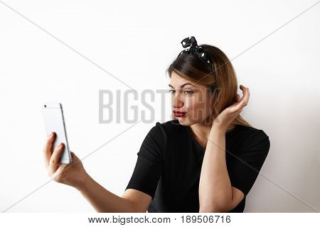 portrait of attractive girl making a duck face posing for selfie holding cell phone. Pretty girl with red lips going crazy and having fun and grimacing posing in studio on white wall background