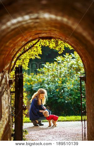 The woman crouched beside the old stone arch in the English garden and strokes her little dog