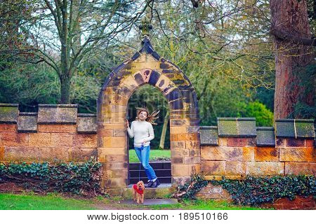 A woman with a small dog accompanies guests standing in an old stone arch in a classic autumn English garden