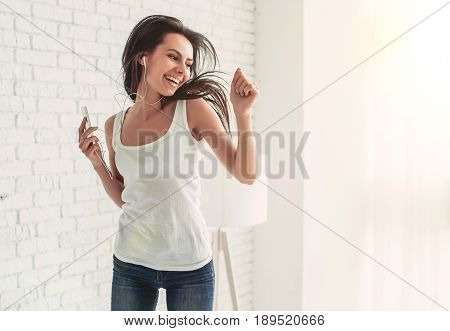 Happy young woman in earphones is listening to music with smart phone dancing and smiling