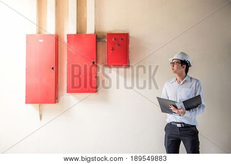 Young Asian Electrical Engineer holding files while wearing a personal protective equipment safety helmet at construction site with red electric control panel. Engineering Maintenance concept.