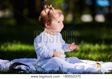 Baby girl in summer dress sitting in the green meadow in the city park. Cute little girl in the beautiful white clothing. Horizontal photo. Sunshiny summer day. Concept of the happy children.