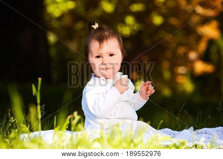 Cheerful baby girl sitting on the green grass in the city park at warm summer day. Cute little girl in the white beauty dress. Horizontal photo. Sunshiny day in outside. Concept of the happy babies.