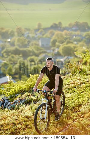 Extreme mountain cyclist riding bike on rocky trail at sunshiny day. Vertical photo. Young sportsman in the black sportwear and with sportive shoes. Beautiful landscape like a background. Concept of the healthy and active lifestyle.