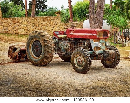 Old fashion antique worn rusted rural red four wheel tractor among green trees. Old agricultural industry machine crawler tractor farm vehicle. Abstract agricultural tractor rusted metal beast