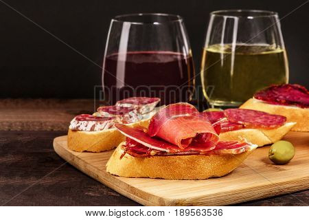 A photo of embutidos tapas, sandwiches with jamon, salchichon, lomo, and other Spanish sausages, with glasses of wine and green olives, on a dark background with a place for text