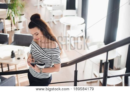 Photo of pretty woman making notes and thinking over a subject while standing in some coffee shop with her laptop near big window