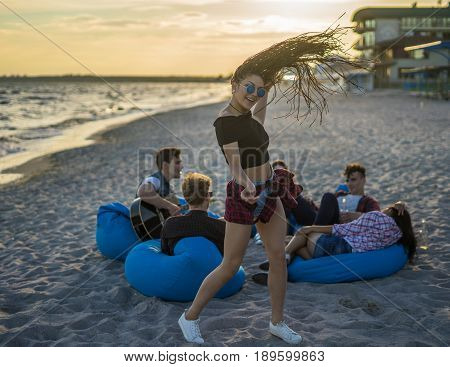 Party on the beach. Sexy girl with zizi cornrows dreadlocks dancing in the foreground, while man is playing on guitar to his friends. They sit on bean bags. Hair up