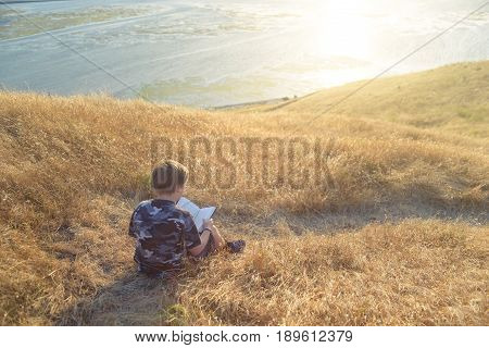 Boy reading book in yellow grass high in the hills, watching beautiful sunset above Bay Area, rear view. Can be used as header.