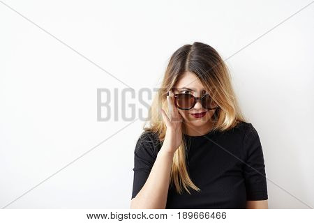 Portrait of smirking Caucasian girl with messy hairstyle wearing balck top and red lips staring from over his sunglasses at the camera. Handsome shopper female posing on white wall background