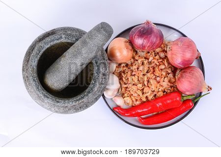 Dried shrimp, fresh red chilies, Bombay onions, shallots, fresh garlic, on a white ceramic plate with blue outer ring line.  Pestle and mortar on a white background.