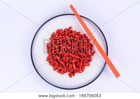 Dried red Goji berries on a white ceramic plate with blue outer ring lines.  An orange chopstick with a white background.