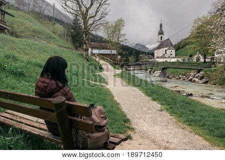 Germany. Ramsau. The Girl Traveler Rests On A Bench Near The  Ramsauer Ache River Overlooking The Pa