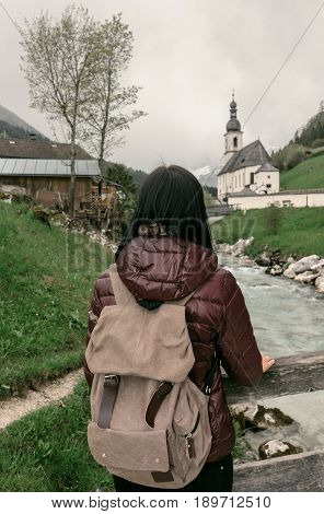 Germany. Ramsau. A Girl Traveler Stands By The Ramsauer Ache River Overlooking The Parish Church Of