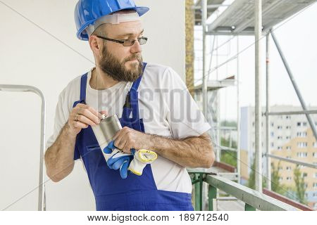 A construction worker in a work outfit and a helmet on the head carefully unscrews an alcohol bottle carefully looking behind him. Work at high altitude. Scaffolding in the background.