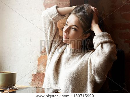 Pretty Caucasian girl joyfully tying her long hair sitting in the coffee shop waiting her friends. Beautyful female student in beige jumper posing in cafe looking with serious expression at the window