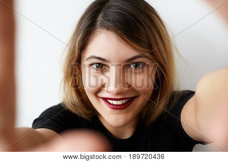 Charming european girl with pretty smile taking selfie on her mobile phone. Young joyful girl holding a smartphone digital camera with her hands and taking a selfie portrait. Casual lifestyle photo.