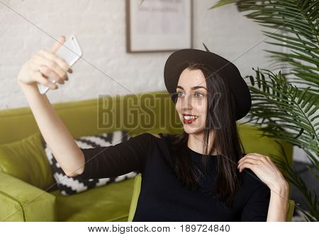Attractive Caucasian girl holding camera and shooting himself sitting in modern interior co-working while waiting her friends. Bright brunette woman is taking fun while making selfie by smart phone