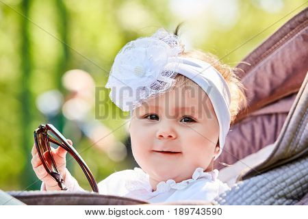 Close-up of the funny baby girl sitting in the stroller and holds sunglasses. Cute little girl in the white dress and with bow on her head. Sunshiny summer day in the city park. Concept of the happy babies.