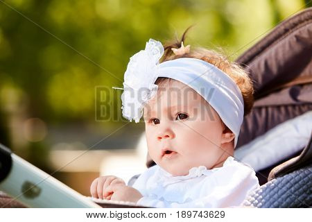 Portrait of the little baby girl sitting in a children's carriage in summer day. Cute baby girl in the white dress and with bow. Horizontal photo and close-up. Warm sunshiny day. Concept of the happy babies.