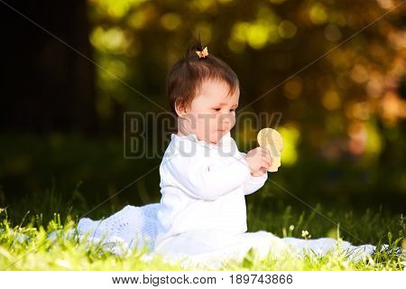 Portrait of happy joyful child sitting on the meadow and holds snack in the park. Cute baby girl in the white dress. Warm sunshiny day. Summer or spring season. Horizontal photo. Concept of the happy babies.