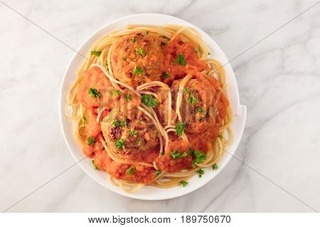 A photo of a plate of meatballs with pasta, tomato sauce, and fresh parsley, shot from above on a white marble texture with a place for text