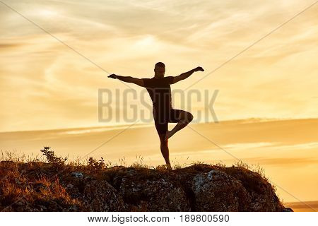 Silhouette of the man practicing yoga on the hill against beautiful sunset. Doing yoga exercises on the nature. Horizontal photo of young sportsman. Beautiful landscape and sky with clouds. Concept of the healthy lifestyle.