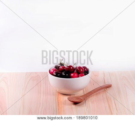 Cherries in white bowl and wooden spoon on white background