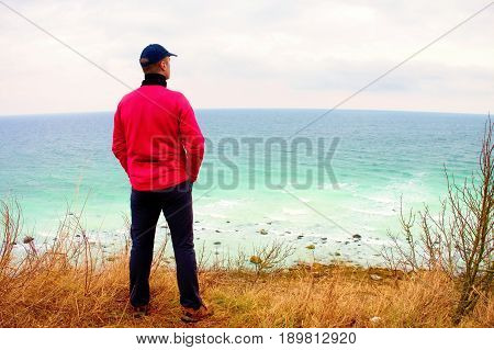 Man In Windcheater Stand On Cliff Above Bay And Looki At Sea Far Horizon. Windy Cold Weather