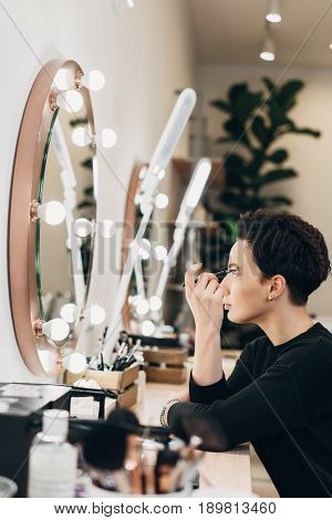 Attractive woman primp at the mirror with lamps in the beauty studio. A happy satisfied customer of make-up and hairstyle service.