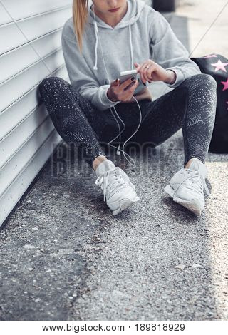 Fitness sporty girl wearing fashion sportswear and sneakers using her smartphone, outdoor sports, urban style. Teen model in swag clothes posing outside.
