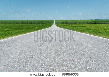 A wide asphalt road between green fields stretching into the blue sky