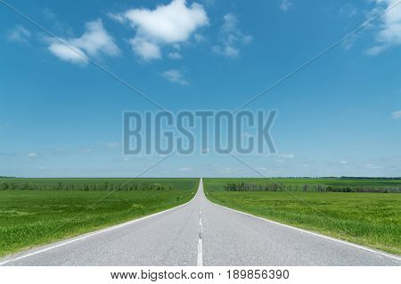 A wide asphalt road between green fields stretching into the blue sky