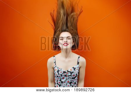 Temperamental Woman with Long Dark Hair Wearing Dress with Flowers is Having Fun in Studio on Orange Background. Hair Style Concept.