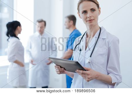Enjoying new approach to ordinary things. Involved skilled smart practitioner standing in the clinic and using digital tablet while her colleagues enjoying conversation in the background