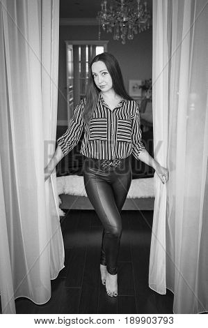 A beautiful brunette with long hair and large expressive brown eyes in a striped blouse and leather leggings stands barefoot between white curtains and smiles a black and white frame