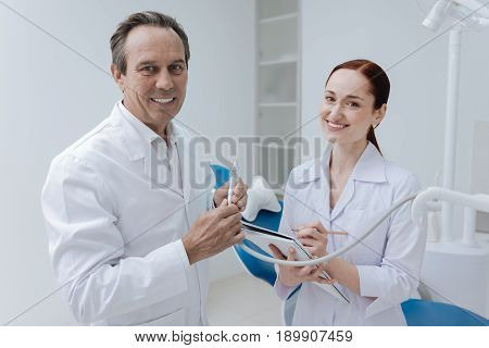 My intern. Positive delighted dentist keeping smile on his face and holding instrument in both hands while standing near his intern