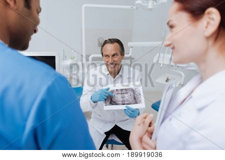 You must know it. Positive delighted stomatologist looking at his intern while sitting on the chair and pointing on X-ray picture